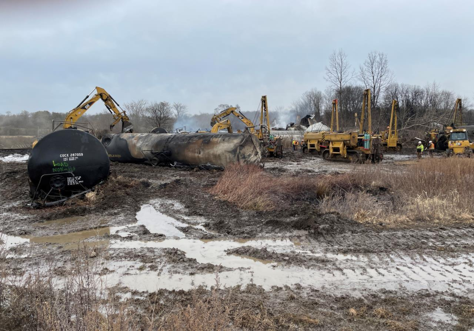 Construction equipment works on derailed trains. There is snow on the ground and bare trees in the background.