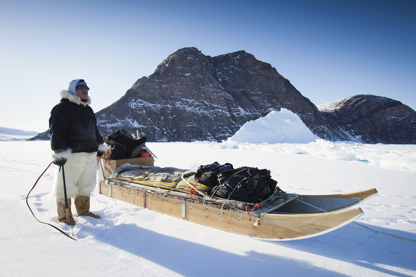 A man wearing fur clothes stands next to his dog sled on the sea ice with a rocky mountain in the background