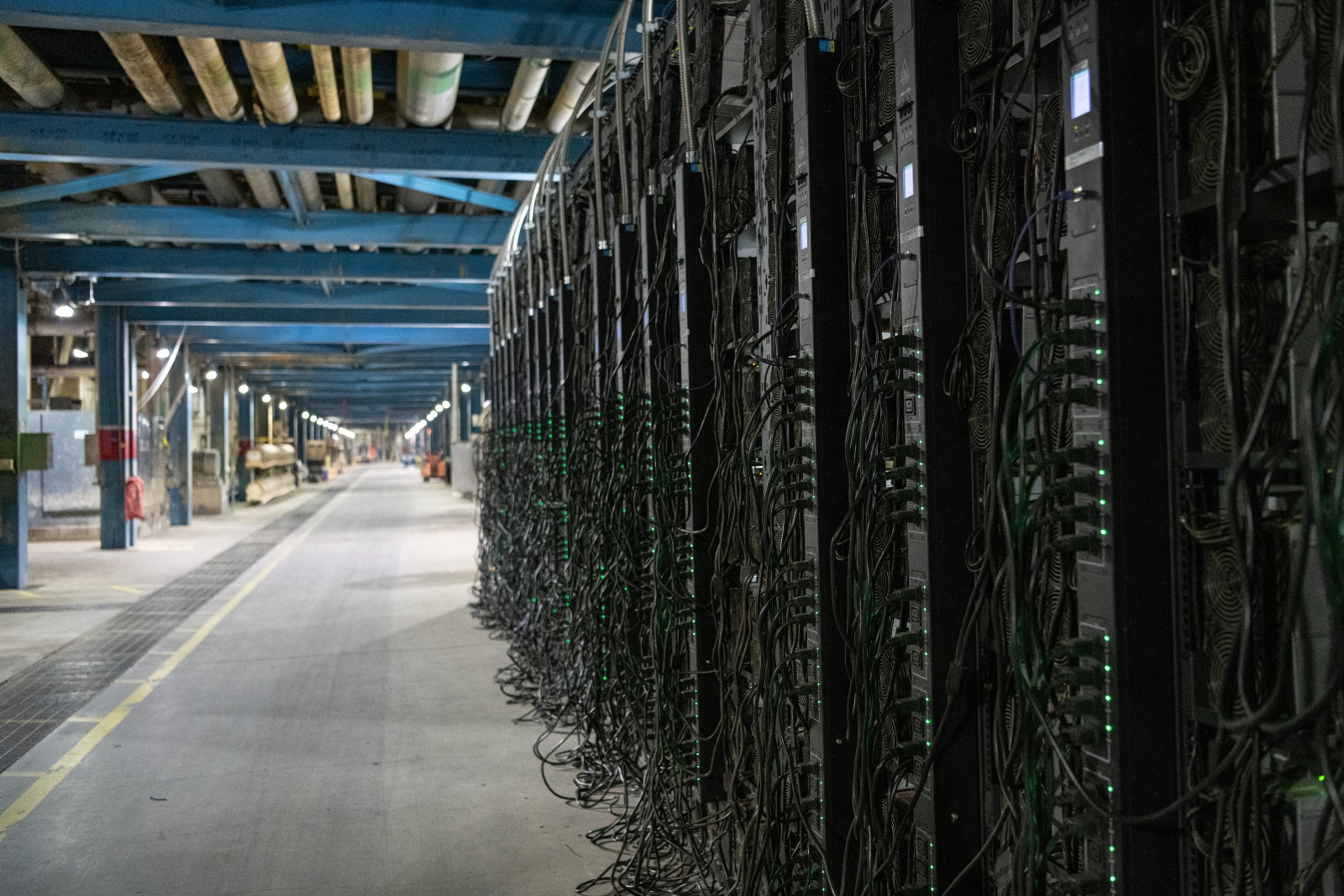 a huge wall of black servers with bright green lights and black cables dangling off of them in an empty hallway.