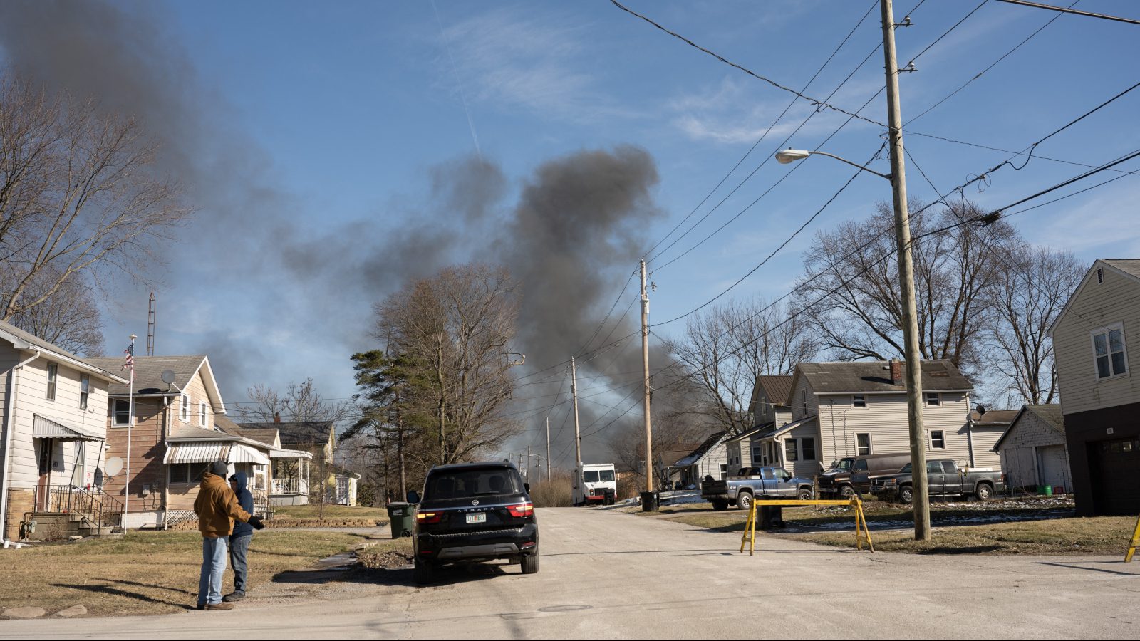 A black smoke cloud can be seen in the distance on a sunny day. The focus shows a residential street with cars, two-story houses, powerlines, and trees without leaves.