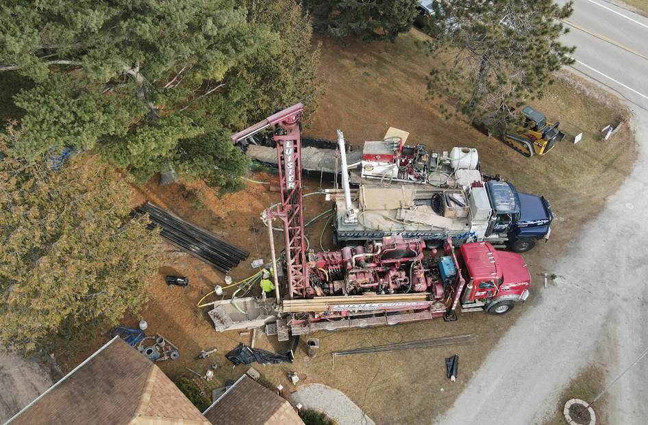 A red and blue contruction truck are seen from above, with green trees surrounding them. Workers are installing a new well in dirt