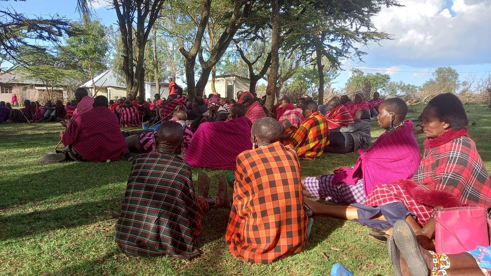 a large group of Maasai sitting on the grass and wearing multicolored blankets