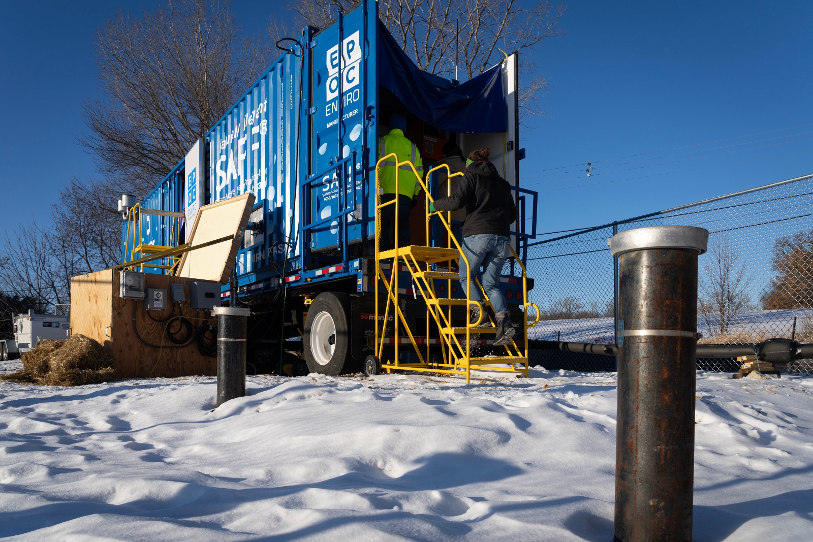 Parked on snowy ground, a blue trailer is open in the back, with two people climbing in through a yellow stepladder
