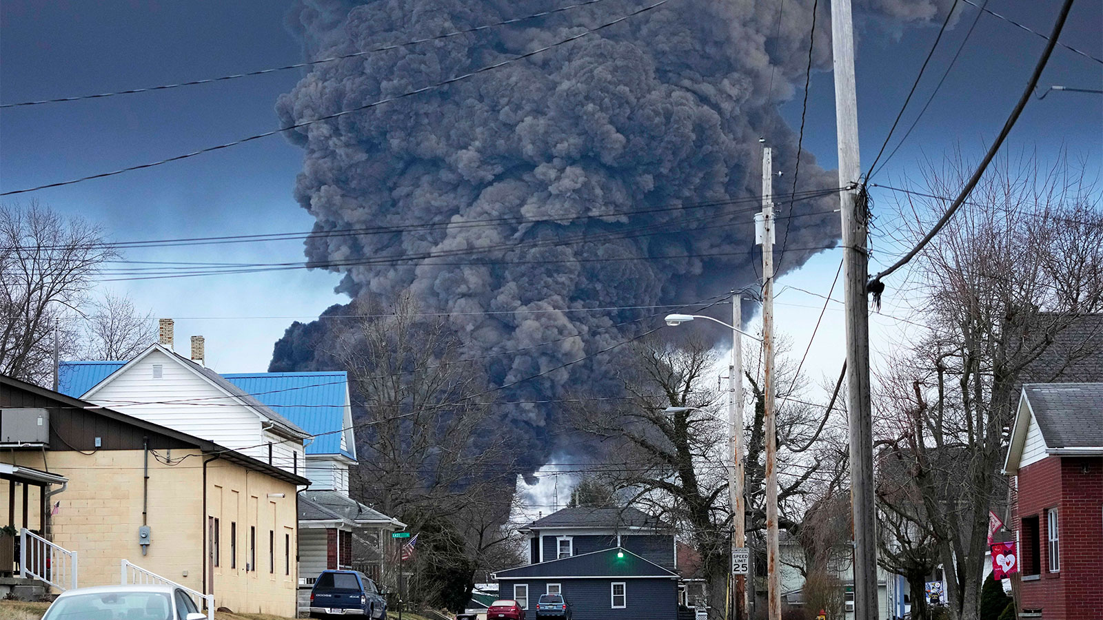 Enormous plume of black smoke billowing up into the sky behind houses on a residential street