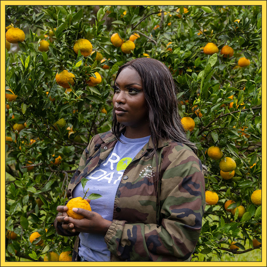 Jonshell Johnson-Whitten stands in front of a satsuma tree, holding a satsuma in her hand