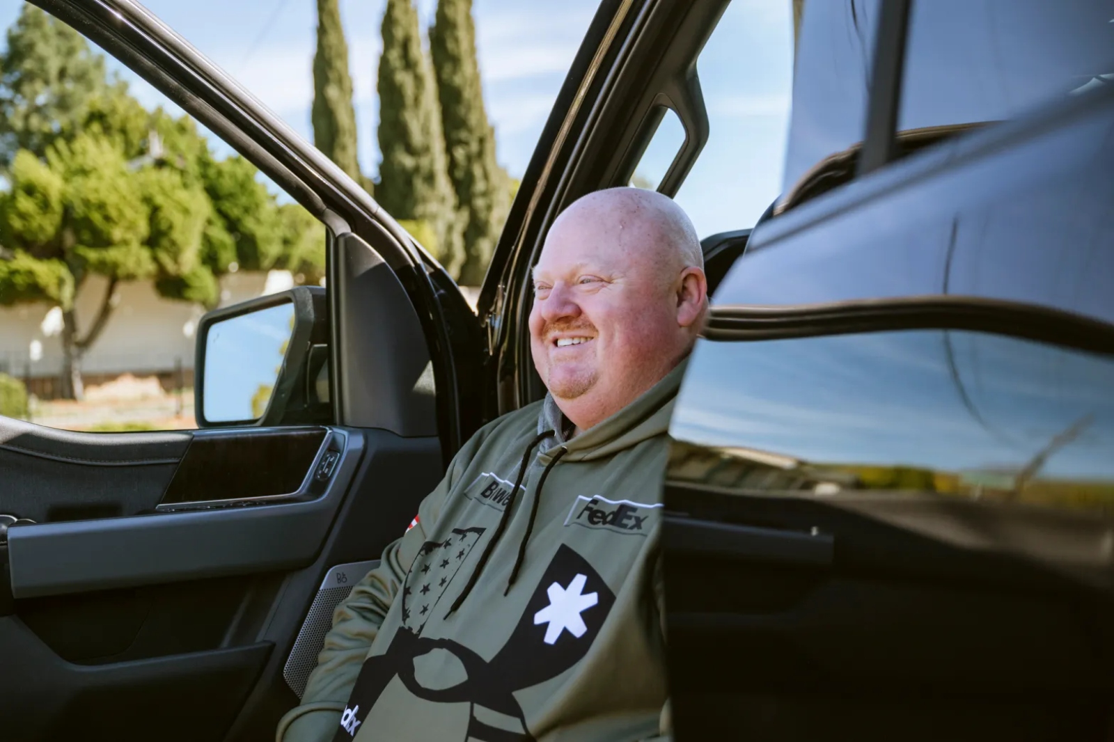 A smiling bald man in a green sweatshirt sits in the front seat of his black truck.