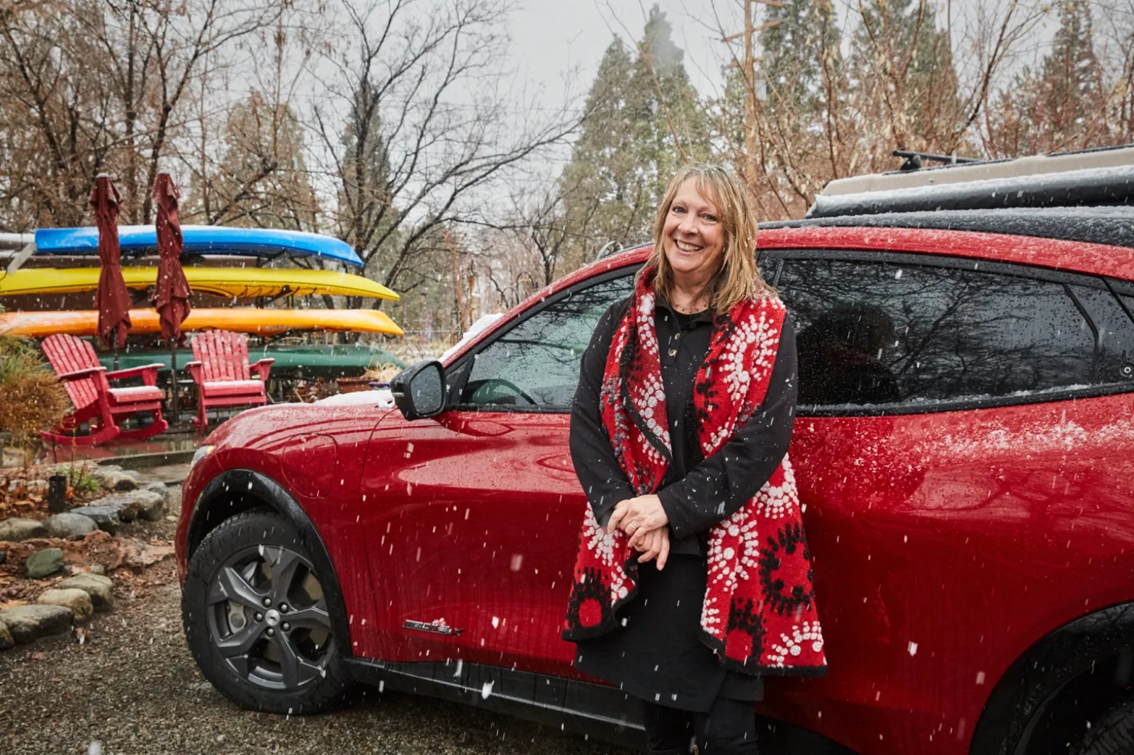 A smiling woman in a red vest stands in front of a red car while it snows.