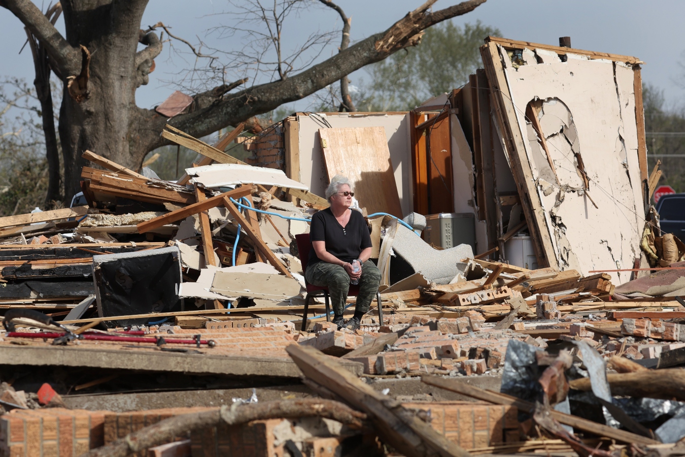 A woman sits on a chair in the middle of a house torn apart by a tornado, a tree with broken branches in the background.