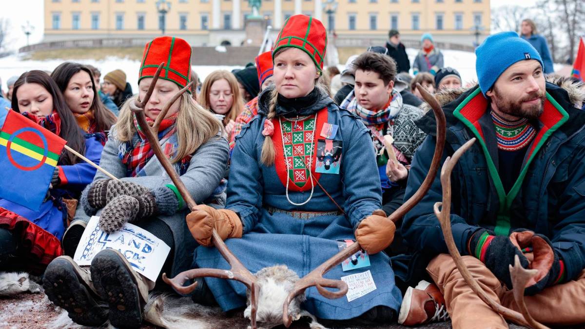 Sámi demonstrators protesting against an illegal wind farm on traditional reindeer grazing lands gather in front of the royal palace in Norway. A young woman holds onto the skull of a reindeer and looks off camera with a stern gaze.