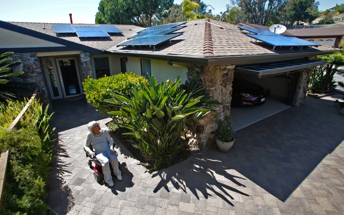 A woman in a wheelchair sits in her sunny patio looking up at the many solar panels on her roof.