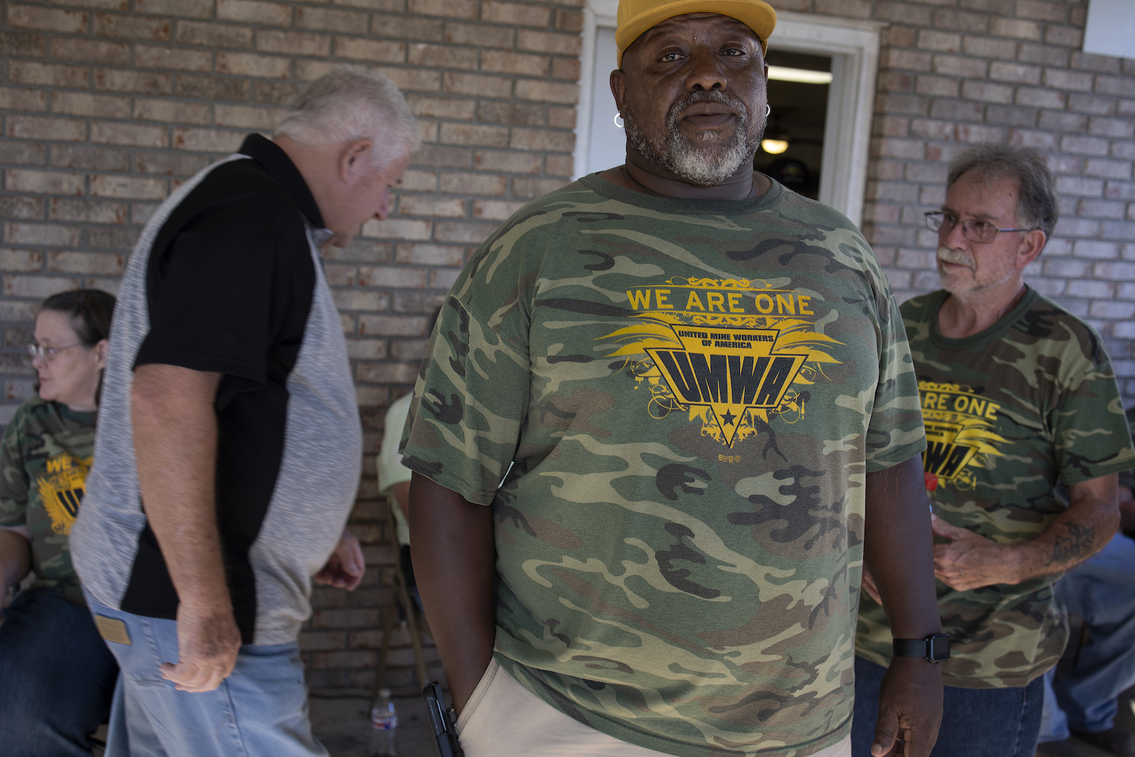 men in camo shirts with a coal miner's union logo stand in front of a brick house