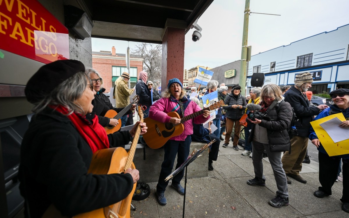 Three older women play guitars and sing, with a crowd around them holding signs and smiling.