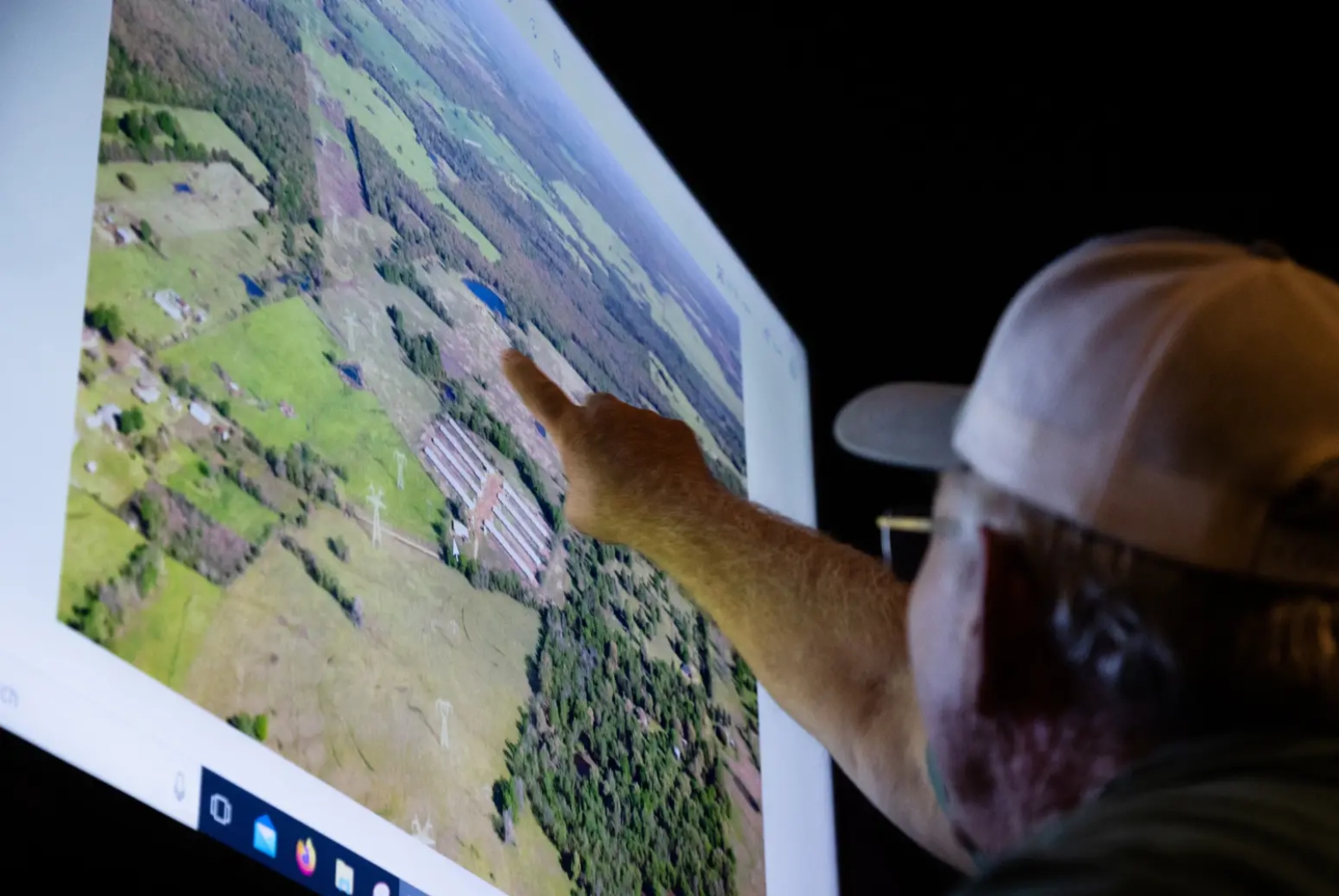 A man in a ball cap and glasses points at an aerial photo.