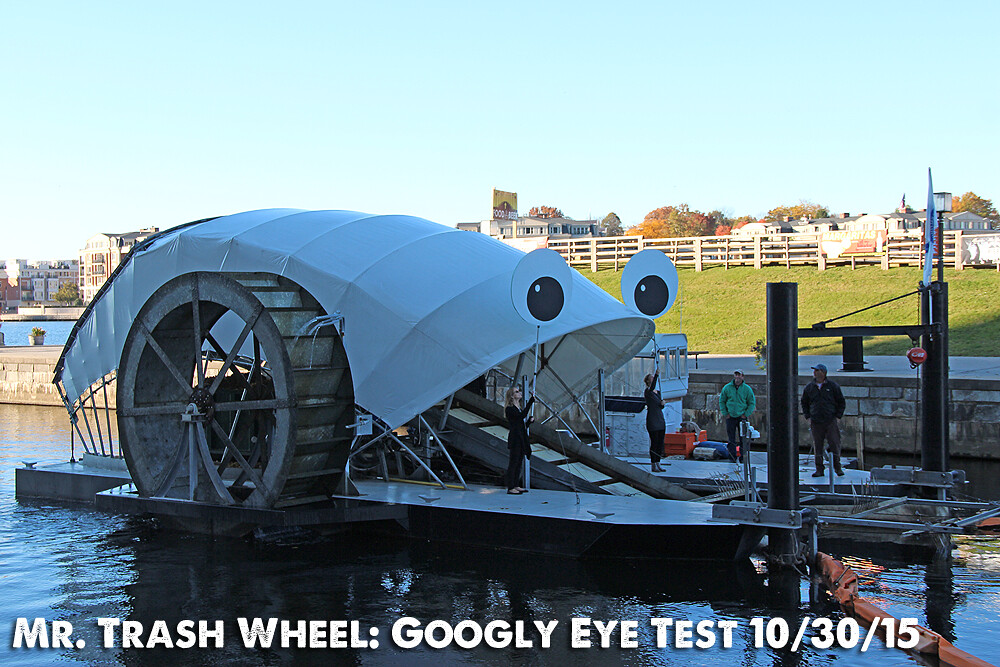 An image of the trash wheel with people standing up the barge, holding up poles bearing large googly eyes.