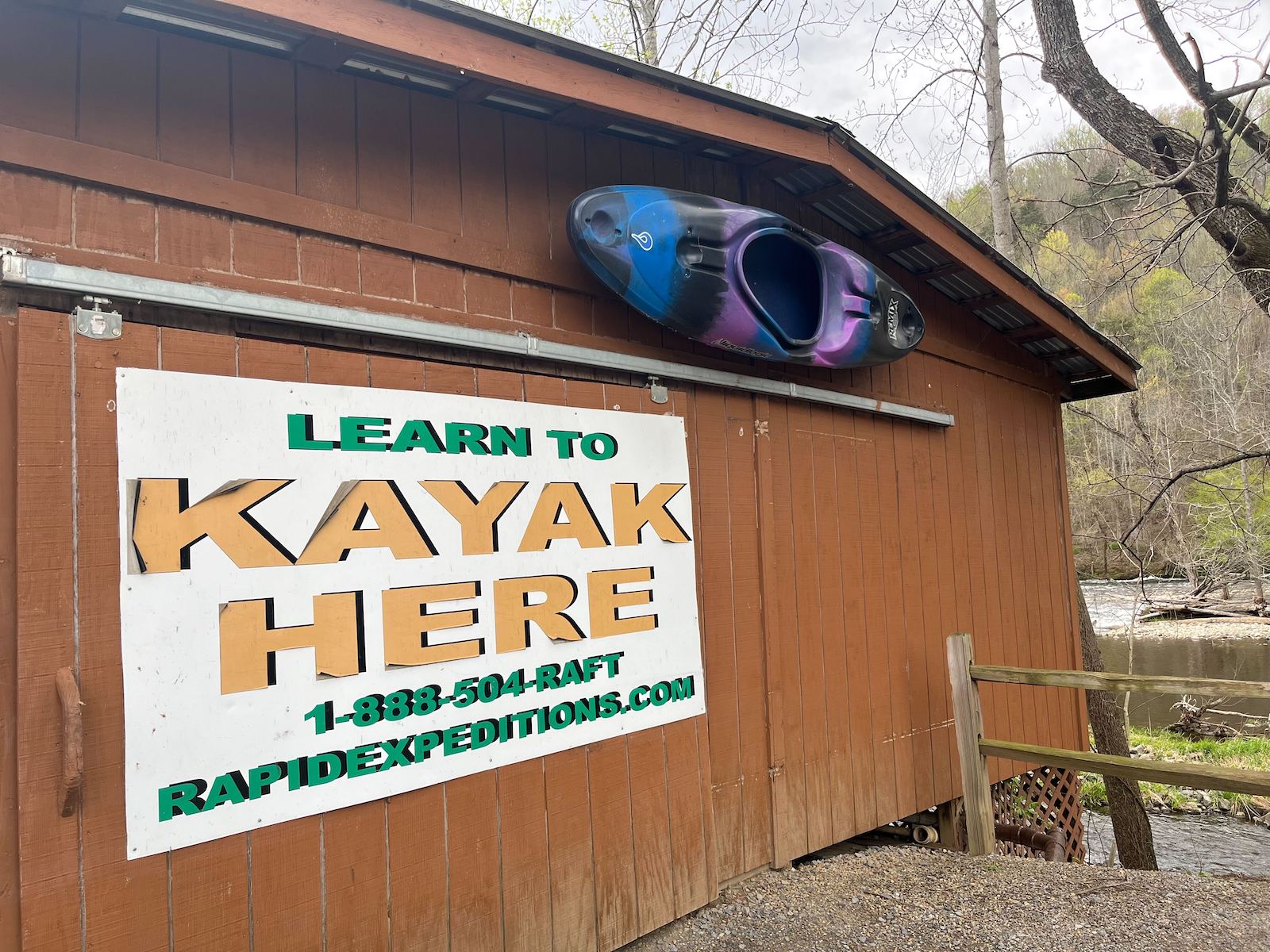 A large white sign that reads “learn to kayak here” on the side of a wood building alongside the Pigeon River in Hartford, Tennessee.