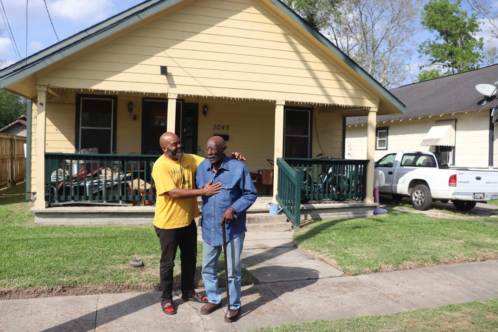 A man in black jeans and a yellow shirt embraces a man in jeans and a blue long-sleeved shirt holding a cane, in front of a yellow house.