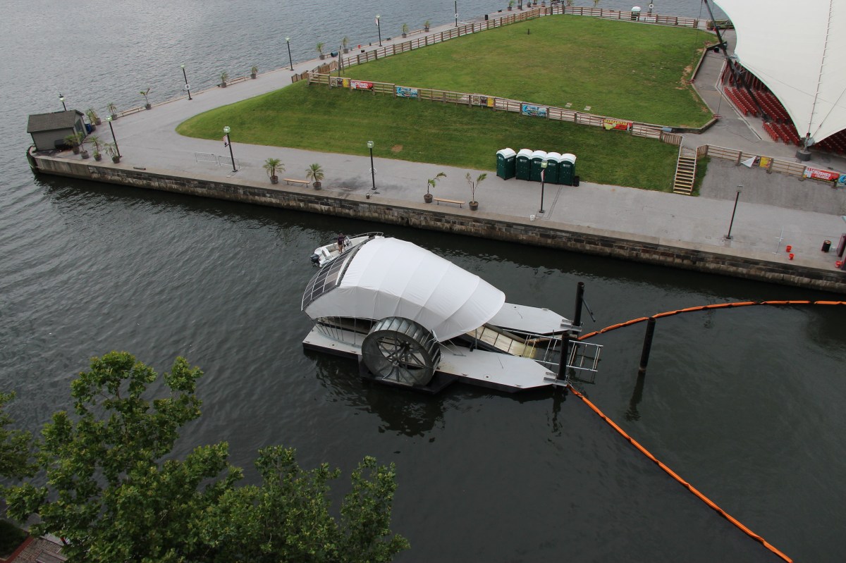 An aerial view of the new trash wheel design on the waterfront, showing solar panels on its back and the lane lines that stretch in front of its mouth.