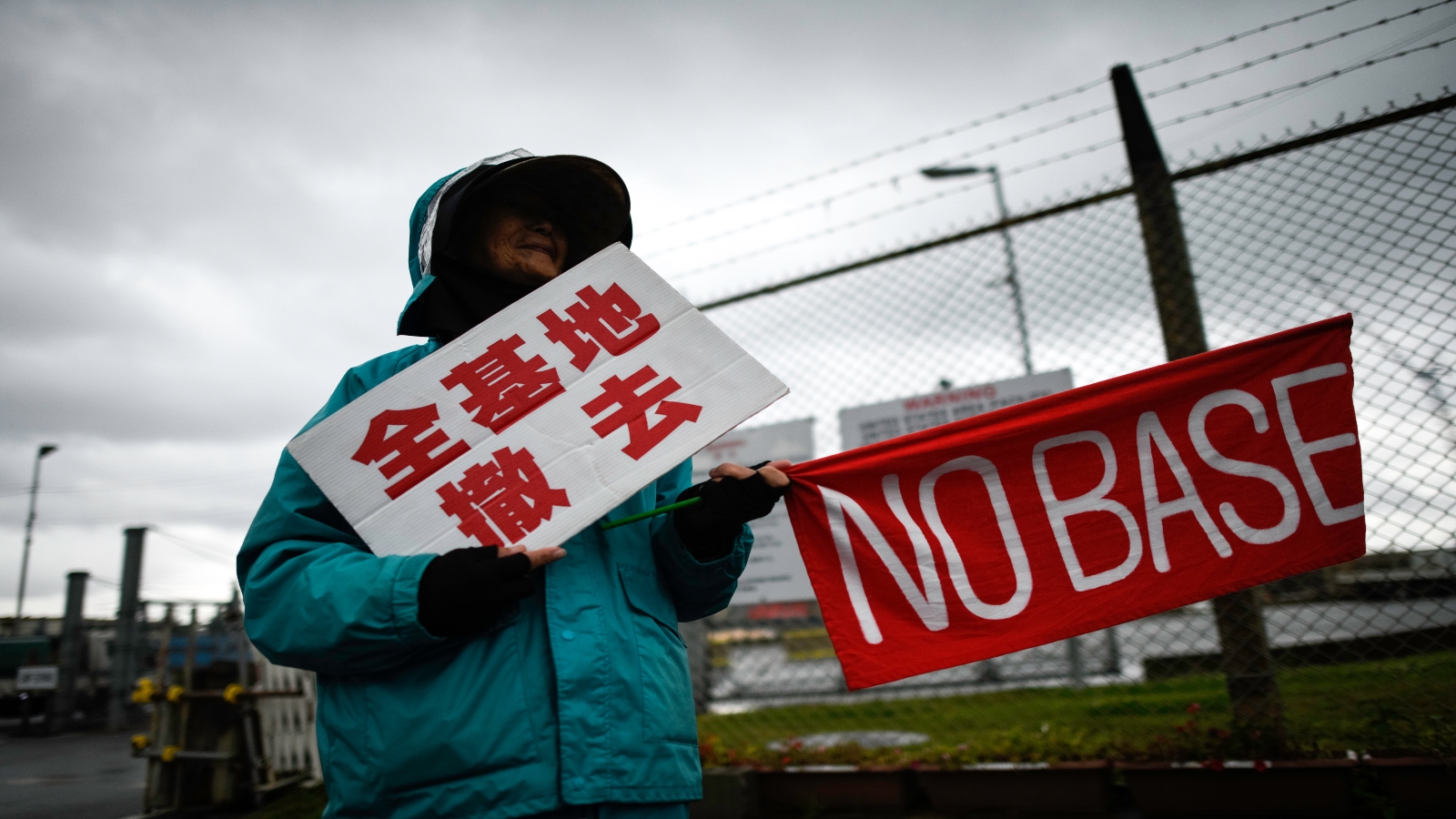 Person in a turquoise jacket standing in front of a barbed wire fence holds two signs, one in Japanese and another that reads 