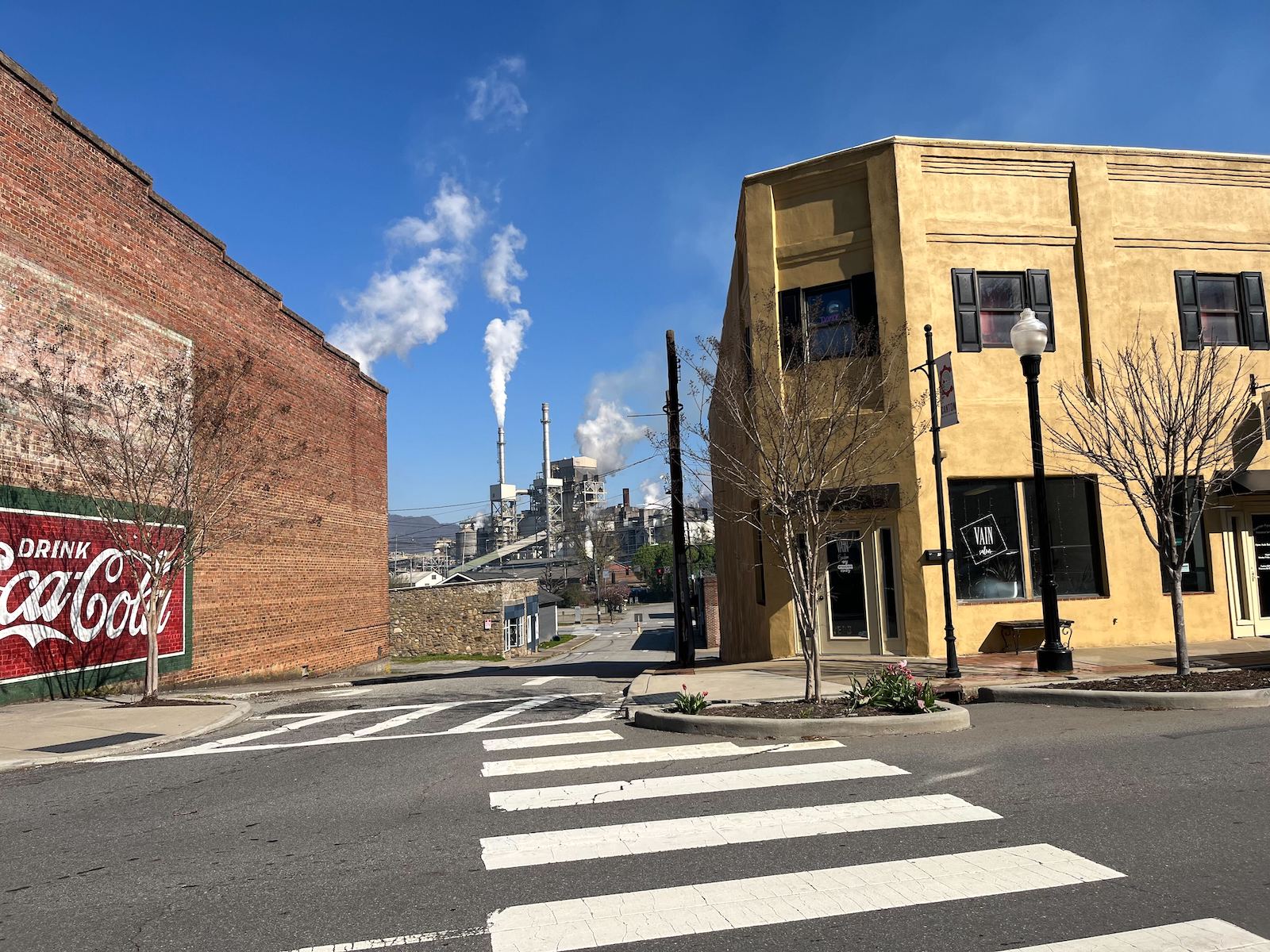 A street running between two buildings in downtown Canton, North Carolina leads toward the paper mill in the distance. Billows of white smoke rise from its stacks.