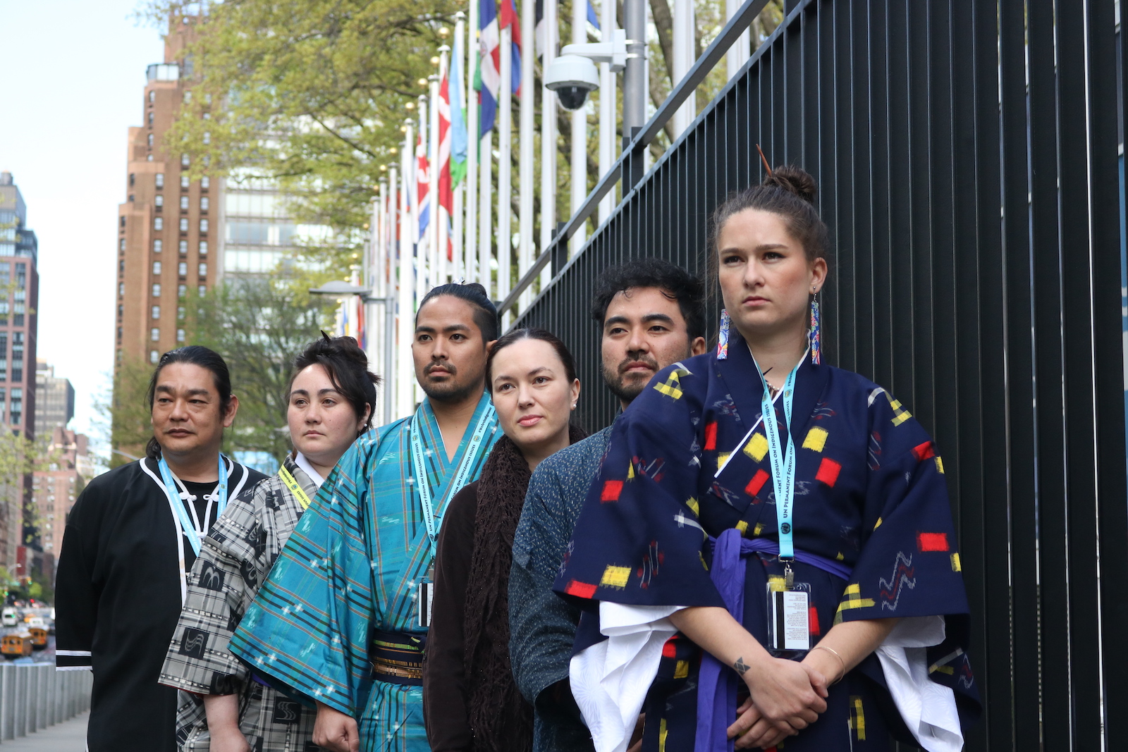 Five people wearing traditional Ryukyuan outfits stand in front of a row of flags at the United Nations, looking off into the distance