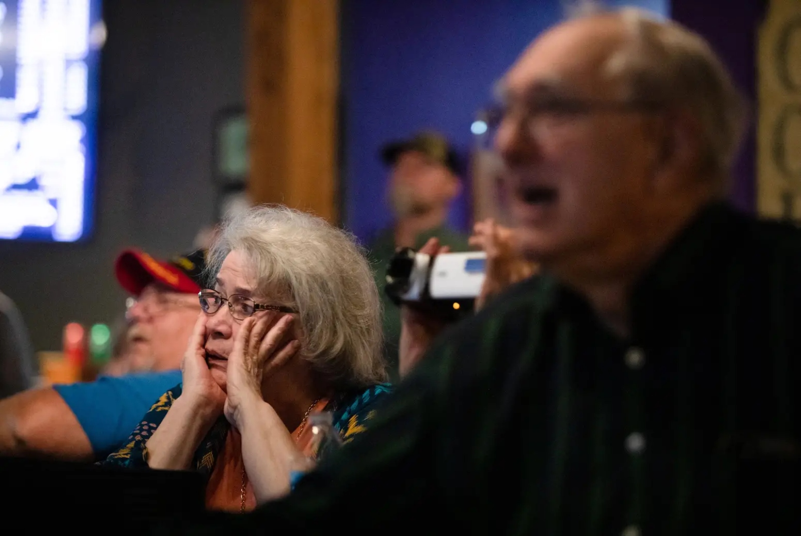 An older woman in glasses grips the sides of her face as a man next to her yells.