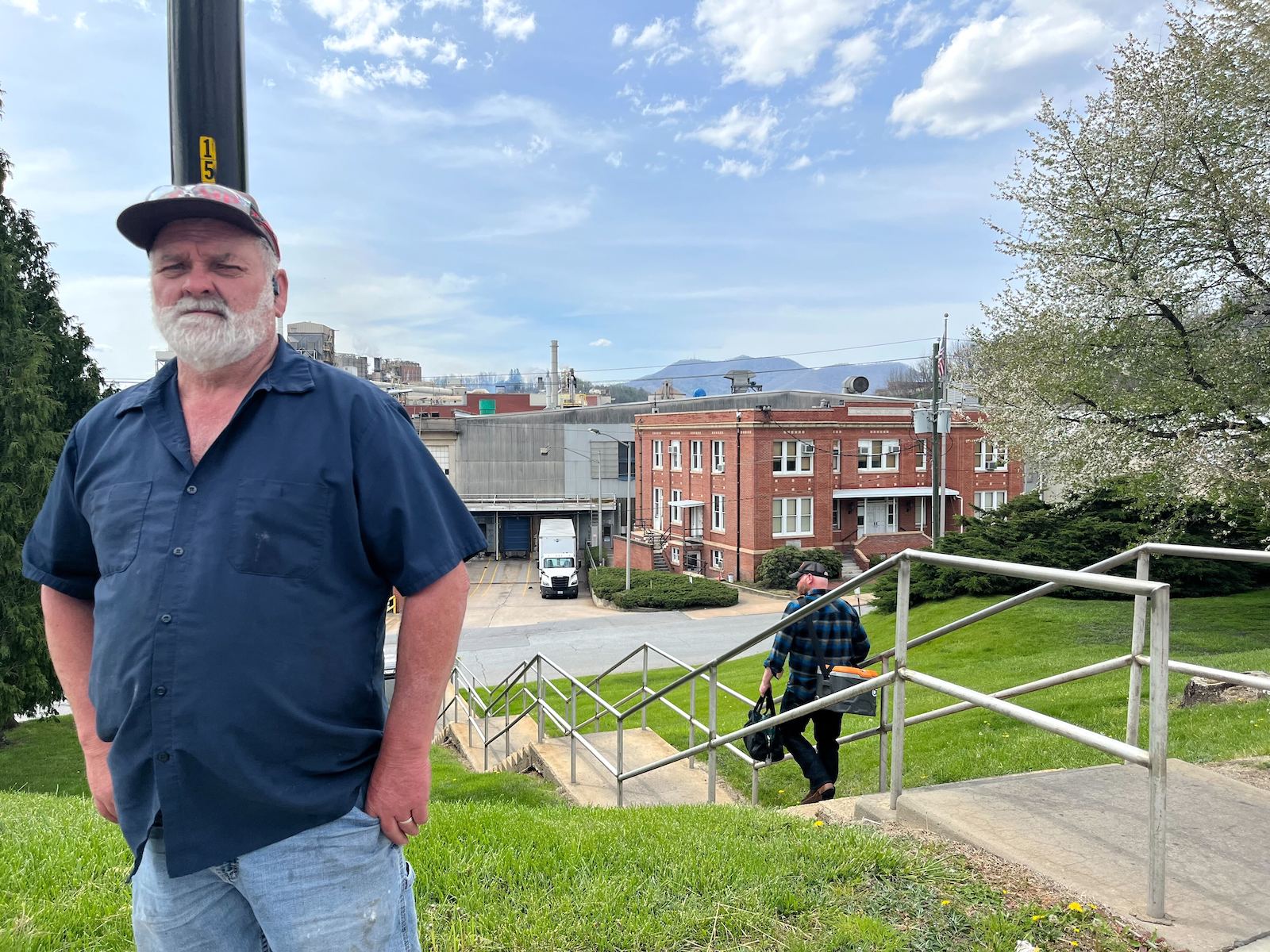 A longtime millworker in Canton, North Carolina, stands on a green lawn alongside a staircase. He is wearing blue jeans, a blue shirt, and a ball cap.