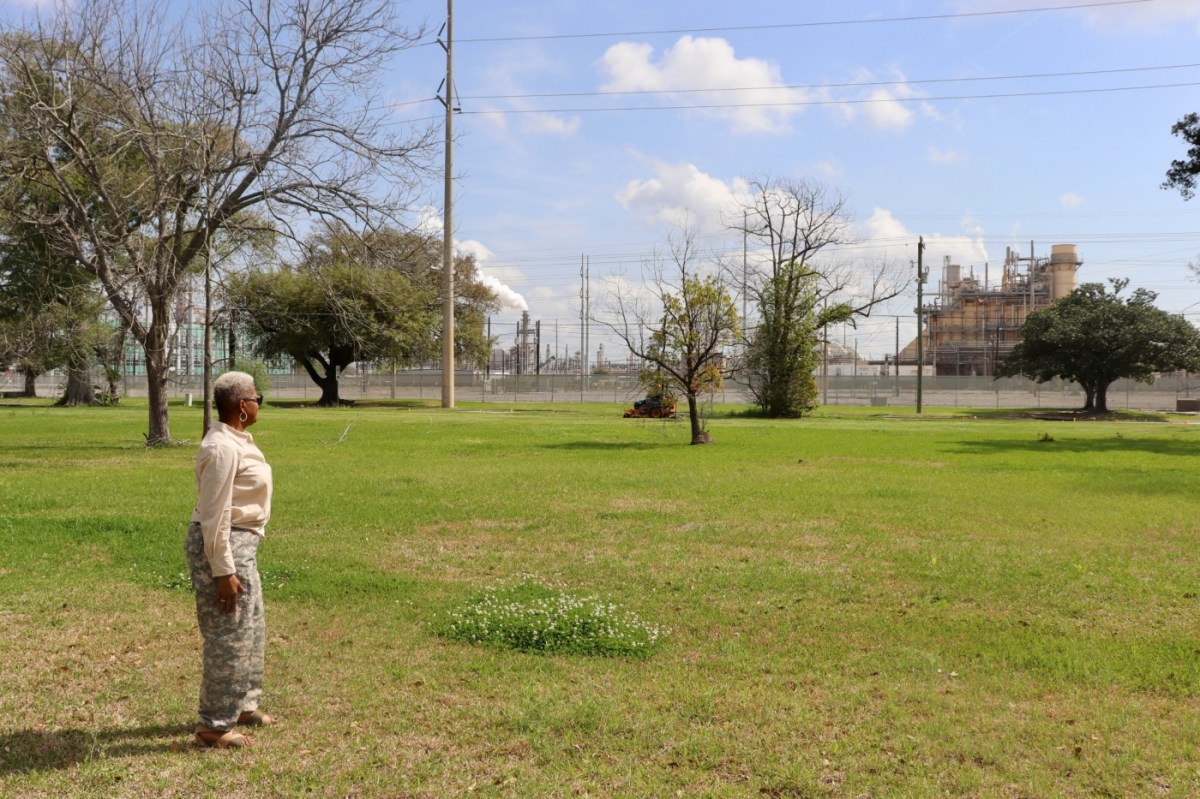 A woman in khakis and a button down shirt and short gray hair stares at an oil plant across a stretch of grass.