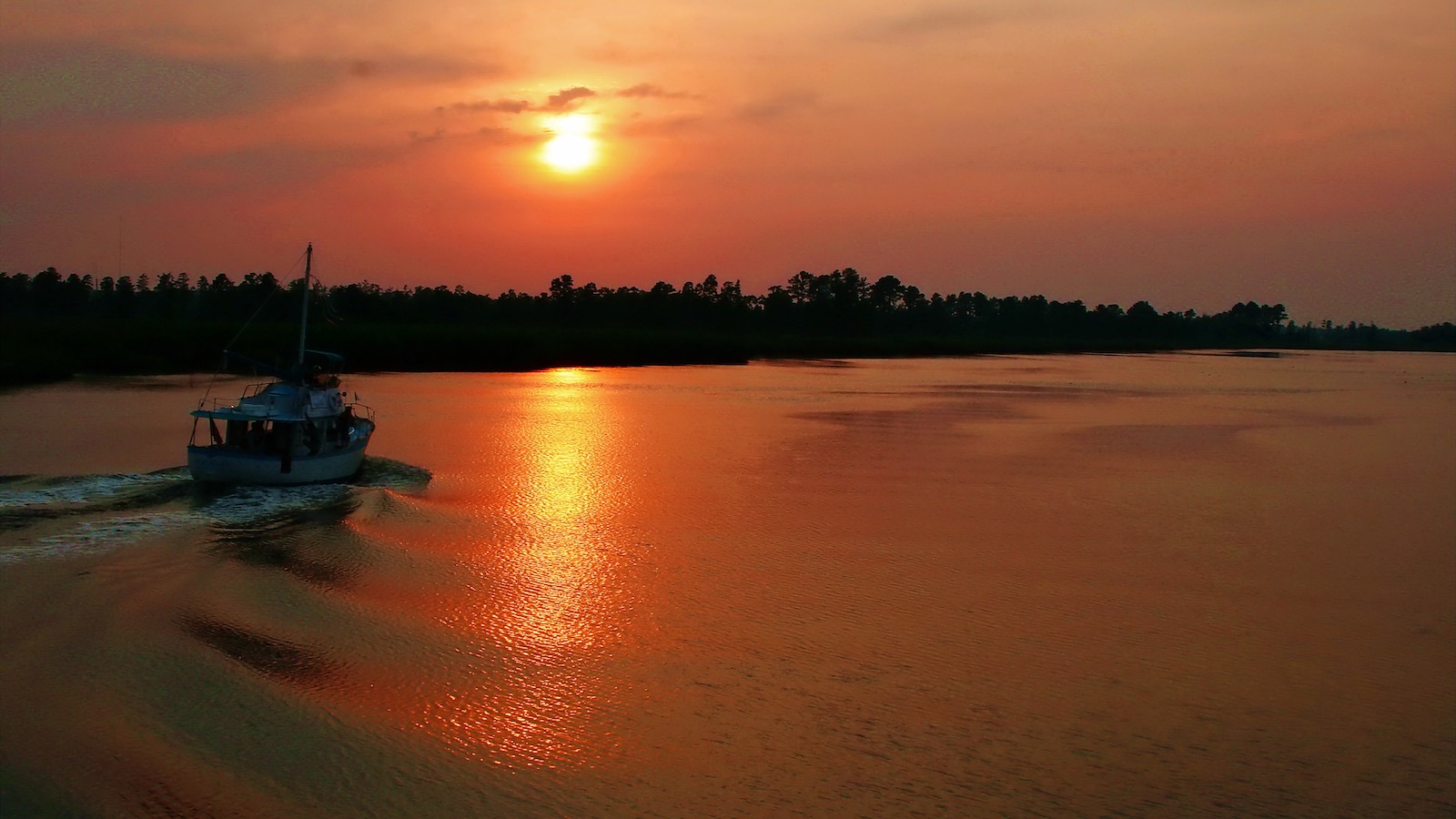 A fishing boat in silhouette heads up the Cape Fear River as sunse casts a golden glow over the water.
