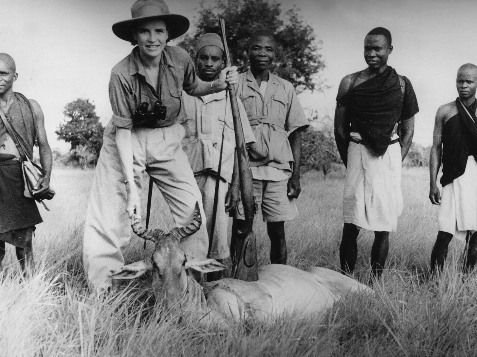 A white woman stands over a dead antelope holding a rifle while behind her stand five African men.