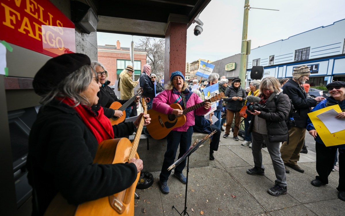 A group of older women holding guitars and singing in front of a smiling crowd.