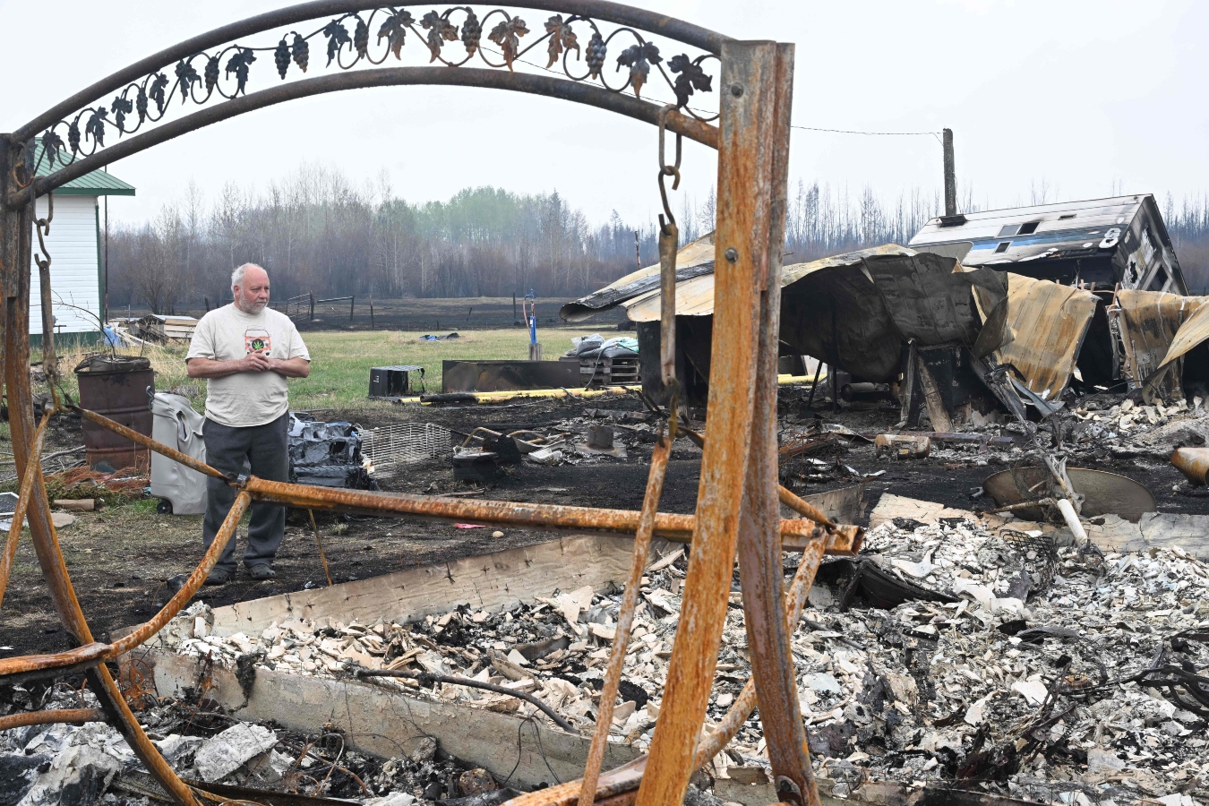 A man stands amid blackened aearth and ashes by a torched porch swing after a wildfire destroyed his home.