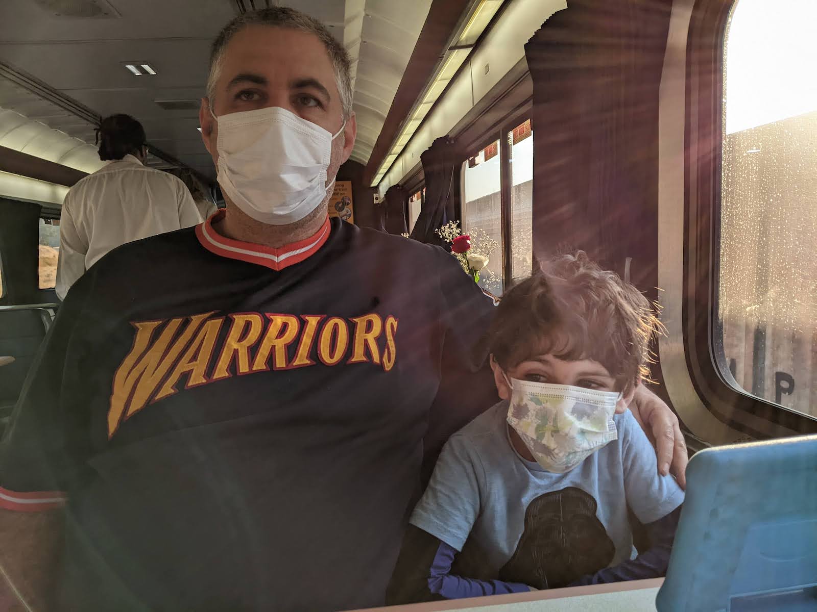 A man in a warrior's shirt and face mask sits in a train seat next to a young boy with a face mask