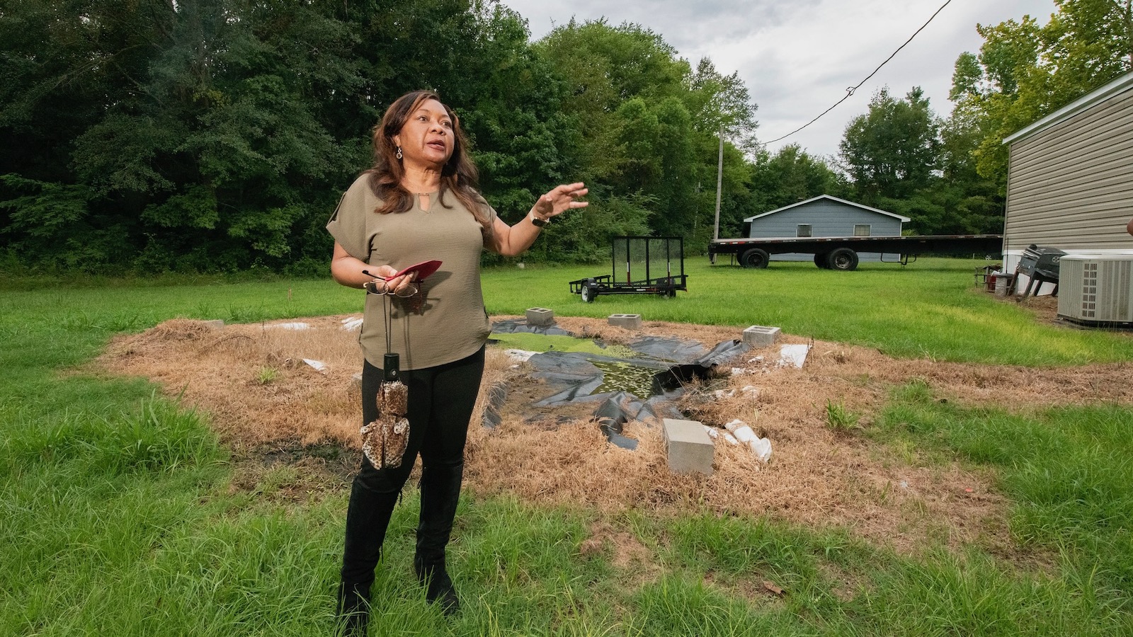 A woman dressed in black pants and an olive green blouse addresses people in the yard of a home in rural Alabama with a failing sanitation septic system.