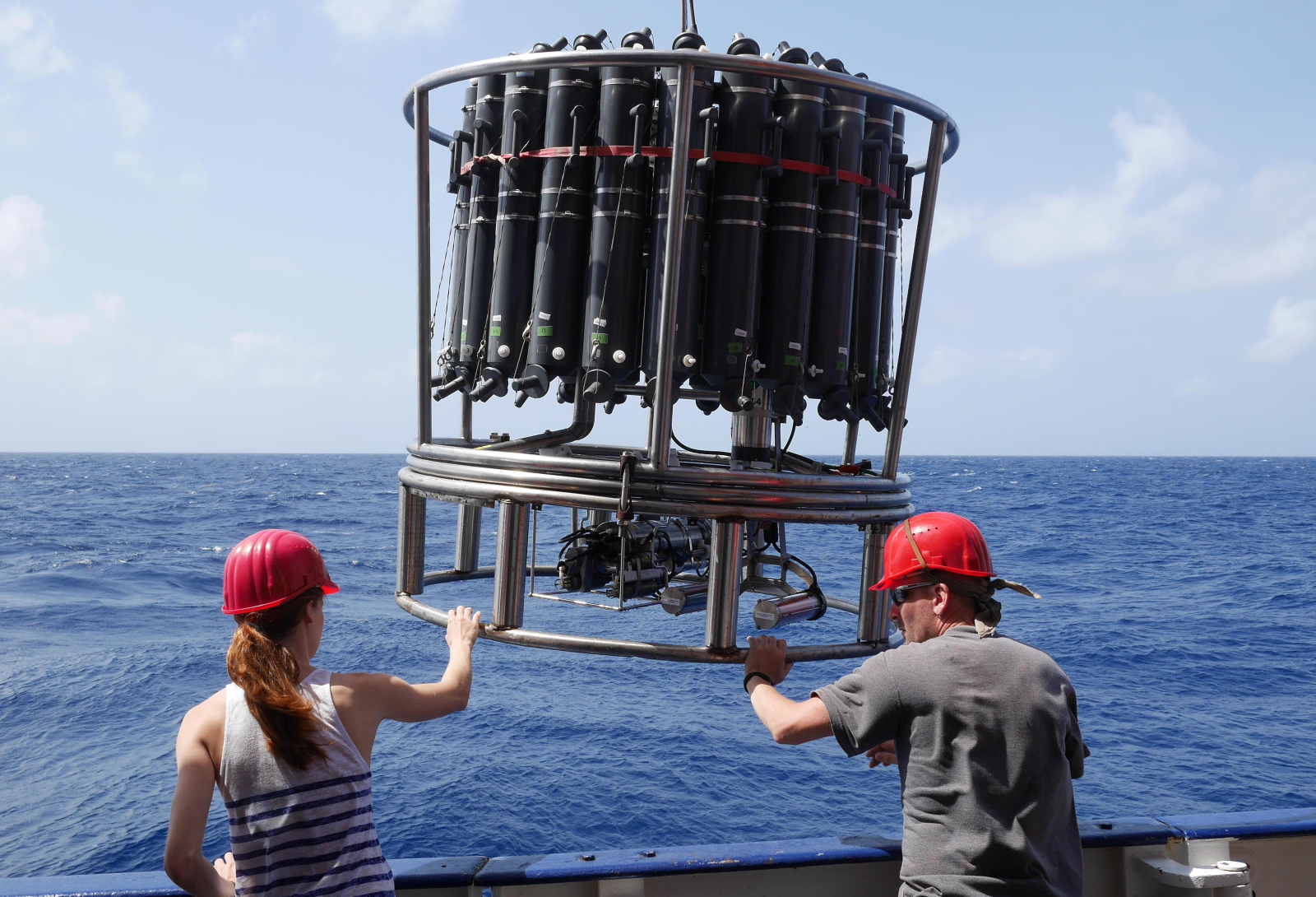 A woman in a tank top and a man in a tee-shirt wear red hard hats and hold on to a metal structure with tanks over the ocean.