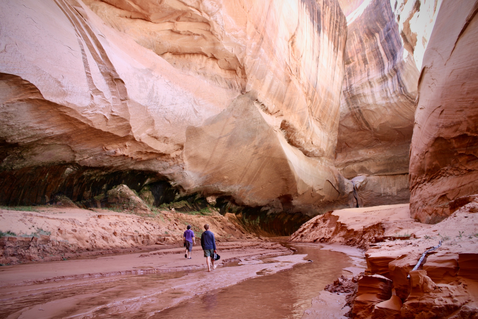 Two people hike through a muddy canyon amid cream-colored rock.