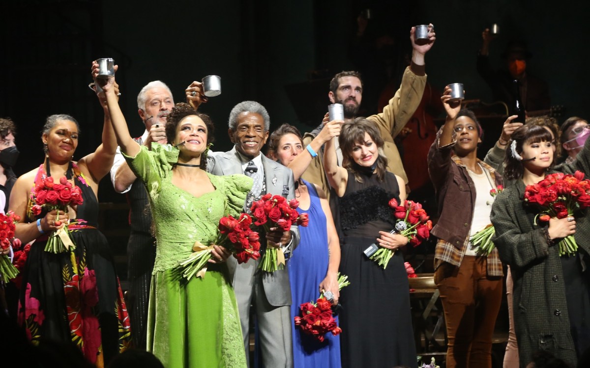 A group of smiling actors holding bouquets of red roses raise up metal cups to their audience.