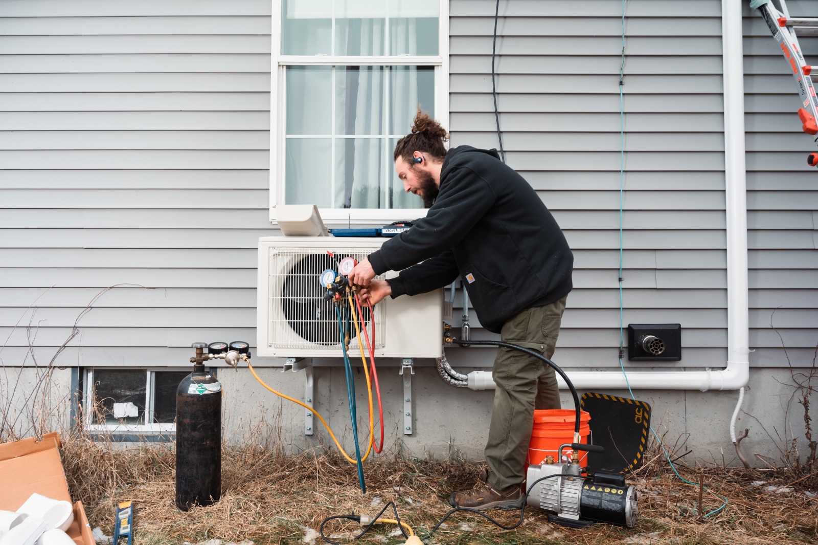 A man with a bun and a black hoodie installs a heat pump on the side of a gray house.