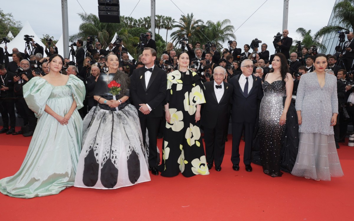 Eight elegantly dressed men and women stand on a red carpet, smiling for photographers.