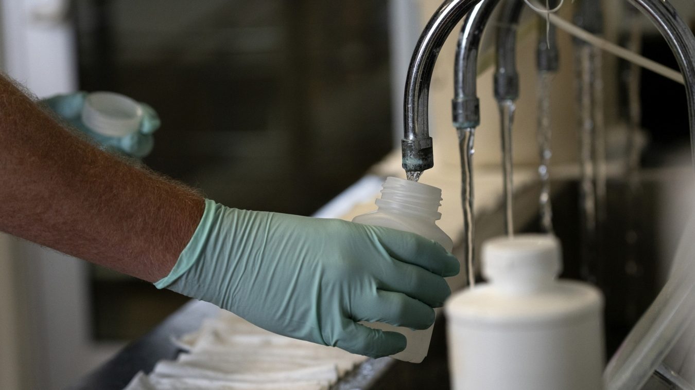 A gloved human hand holds a plastic bottle under a faucet.