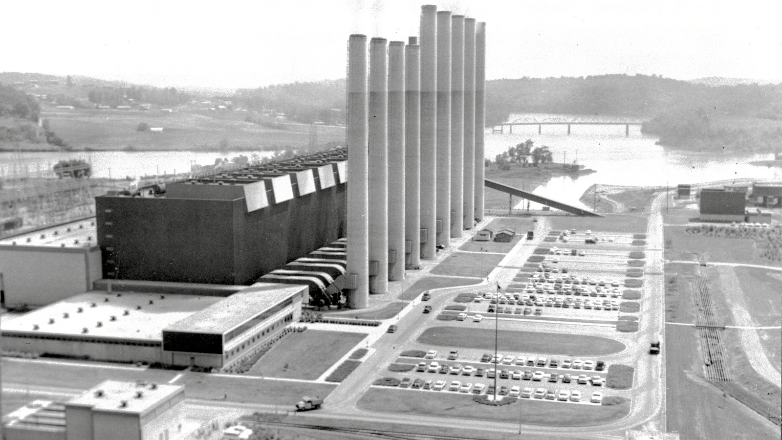 A black-and-white photo shows the main building and nine towering smokestacks of the Kingston Fossil Plant on the banks of the Clinch River near Kingston, Tennessee.