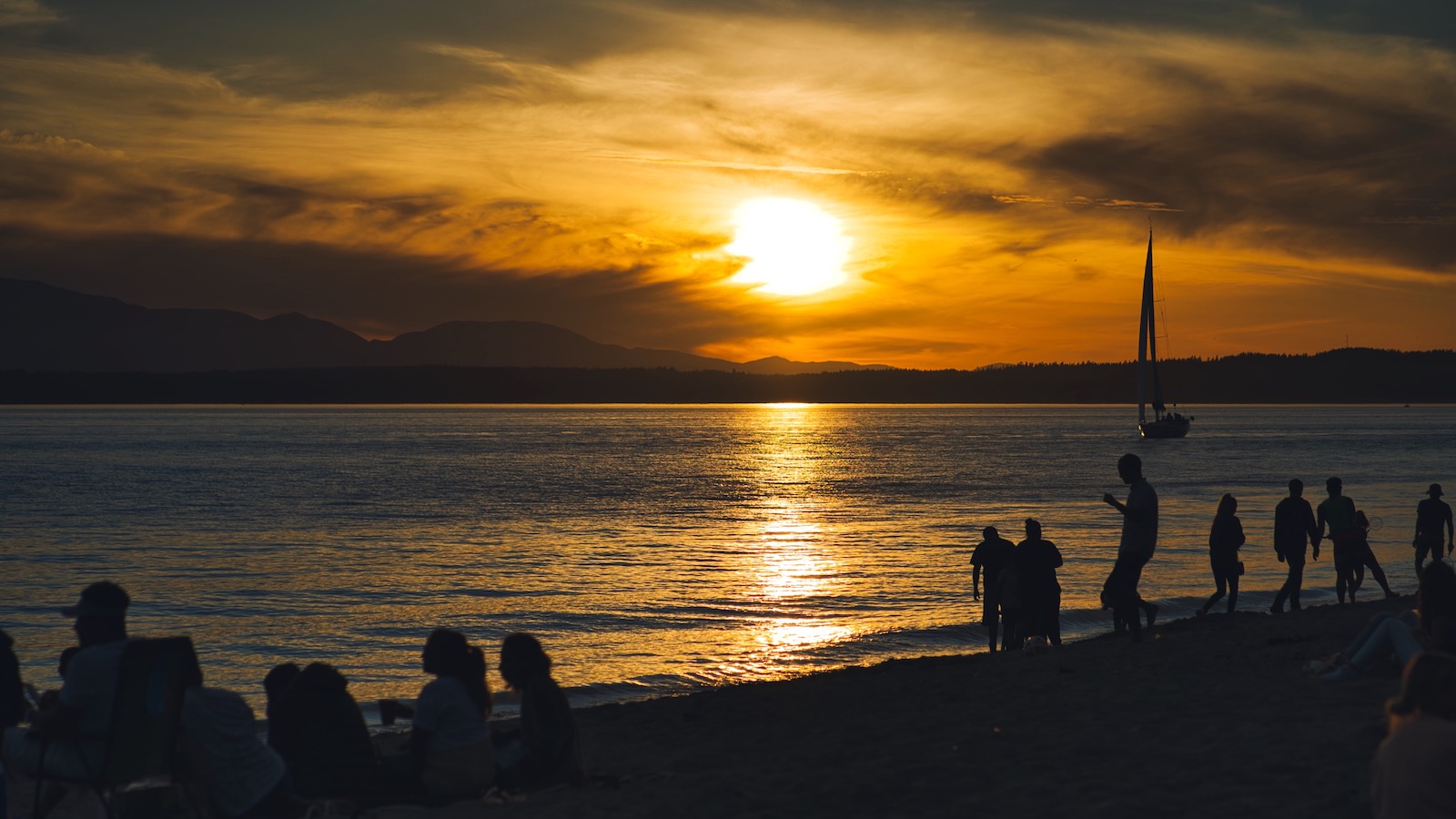 People on a beach in Seattle are seen in silhouette as the sun sets over the mountains in the background.