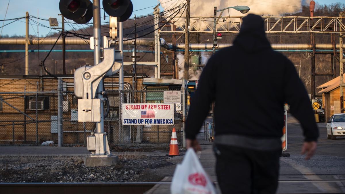 A man seen in silhouette walks toward a steel mill, where a banner reads "Stand up for steel Stand up for America.