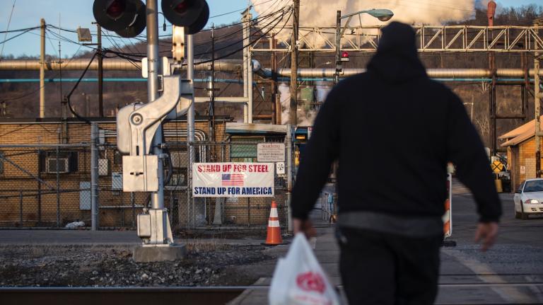 A man seen in silhouette walks toward a steel mill, where a banner reads 
