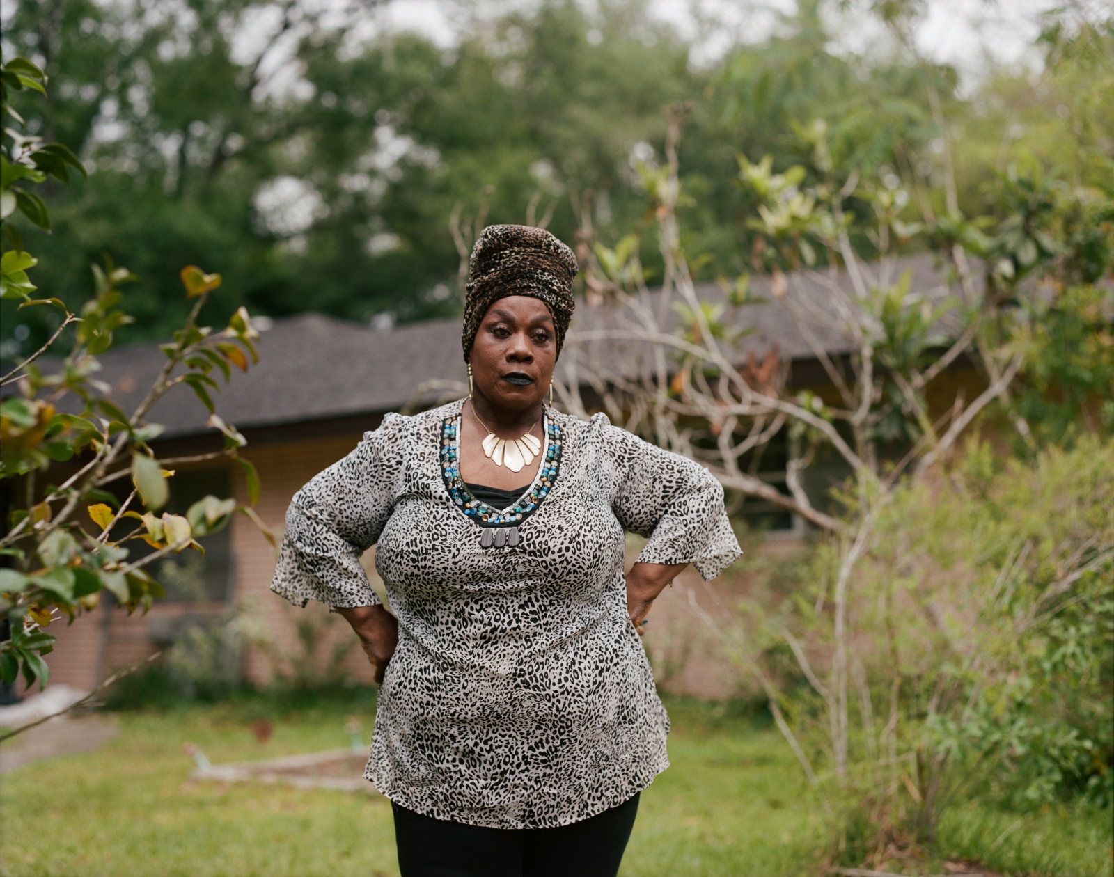 A woman with a large necklace and head wrap stands with her hands on her hips in front of a house.