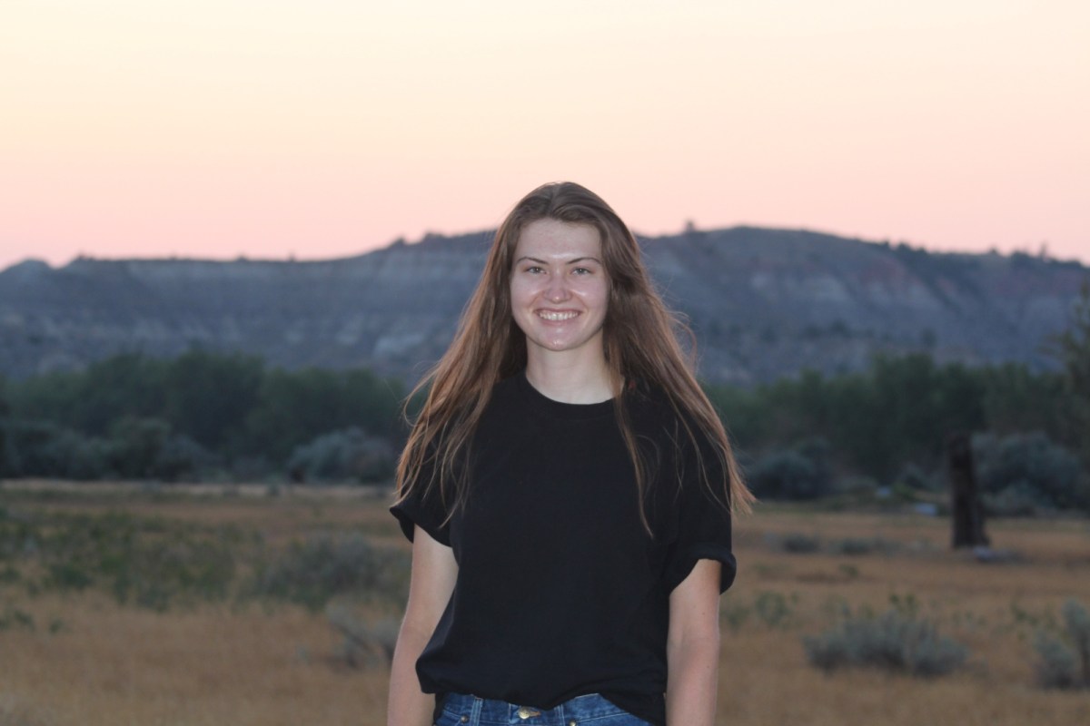 A teenager in a black tee-shirt with long hair stands in a field at sunset.