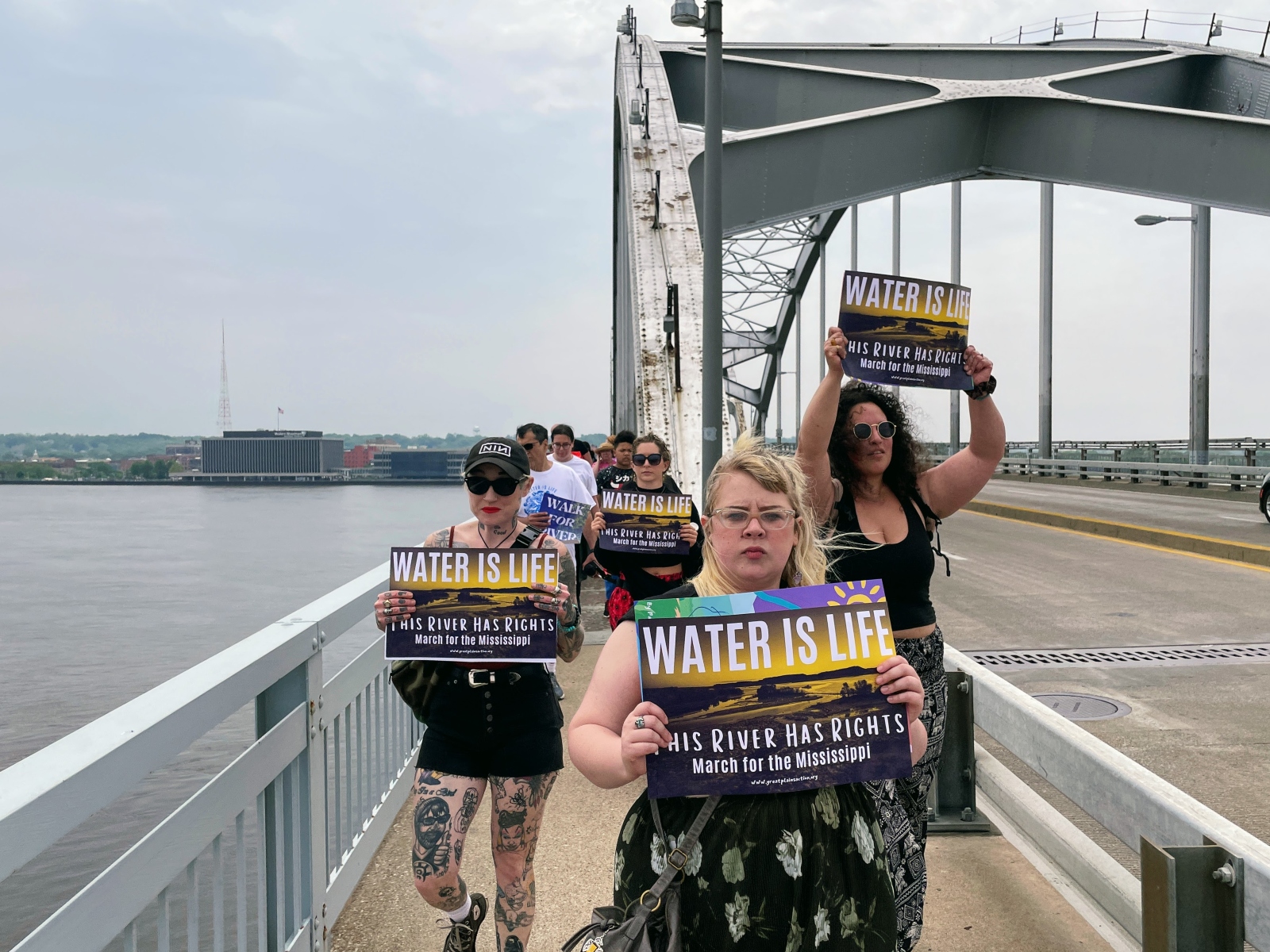 A group of young people walks on a bridge over a river with "water is life" signs.