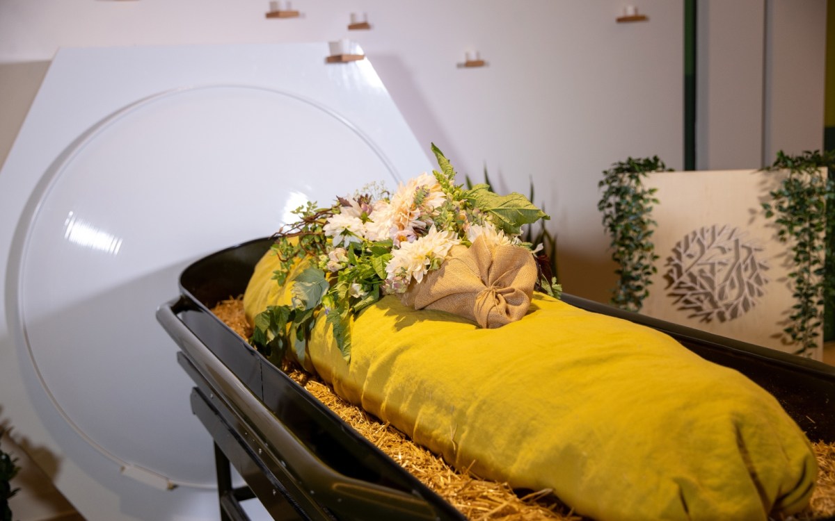 A casket holds wood chips and a shrouded form with a bundle of flowers placed on top.
