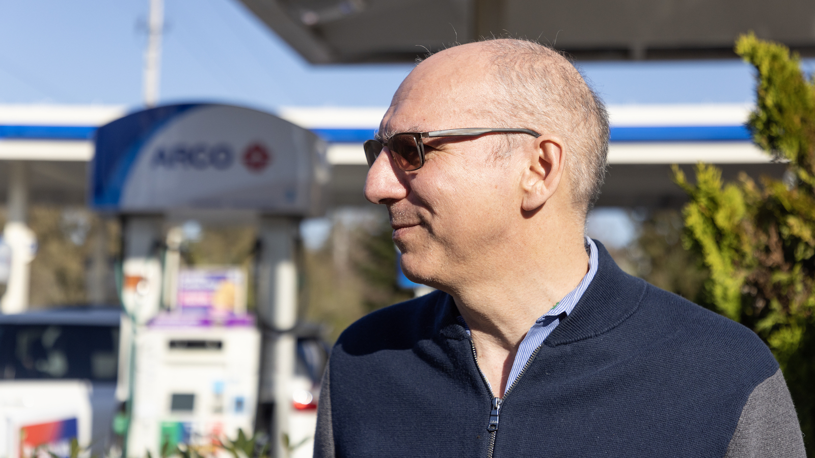 a bald man with sunglasses in front of an ARCO station