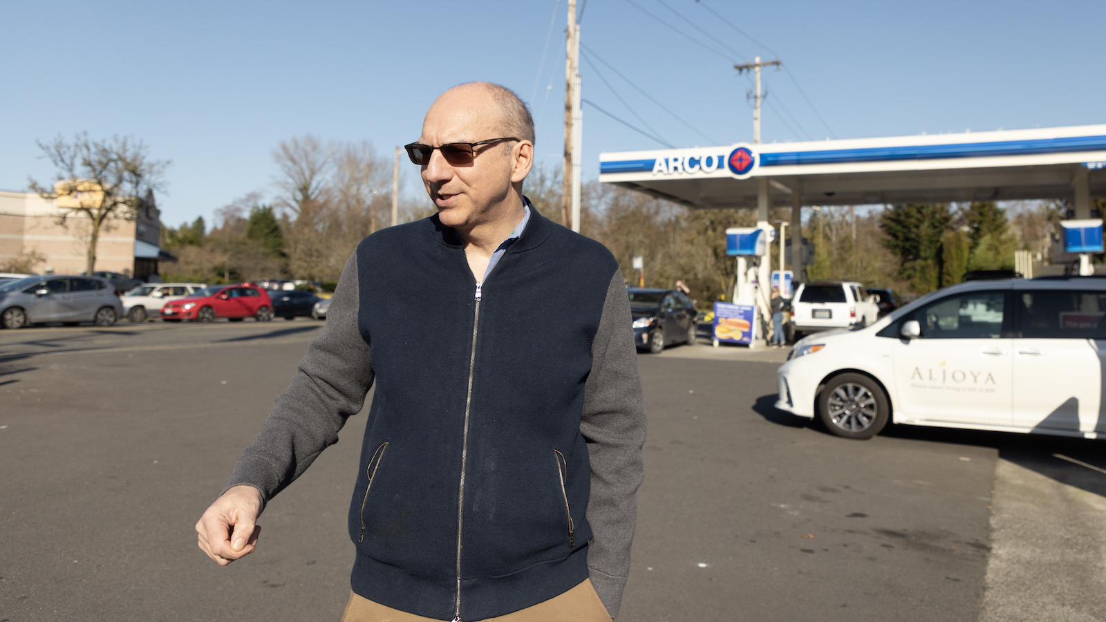 a man in a jacket and sunglasses walks in front of a gas station