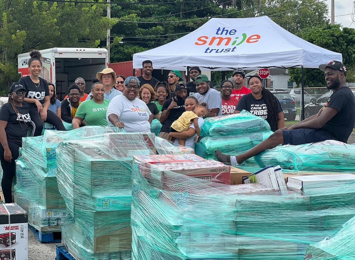 Gunder and a large group of volunteers stand smiling around plastic pallets piled high with wrapped boxes of supplies. 